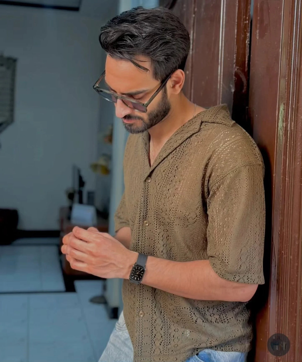 Man wearing a brown patterned shirt and glasses, leaning against a wooden door.